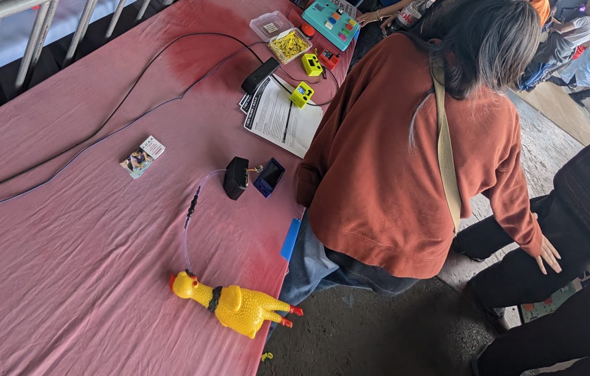 A table of interactive assistive devices: button boards, a sip-and-puff sensor and a yellow rubber chicken, with a visitor leaning in to try them
