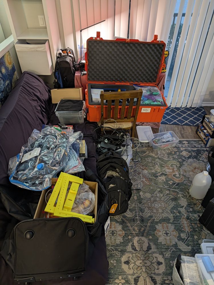 Living-room floor covered in packing: an orange hard case of devices, bags of parts, a yellow printed stand, backpacks and cables